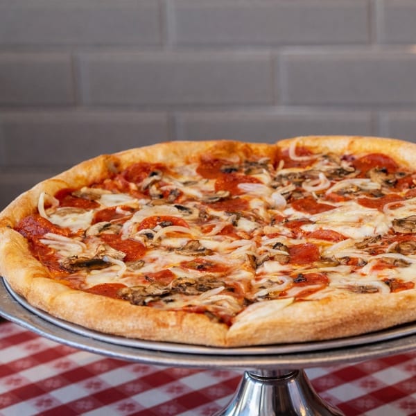 A whole pepperoni, mushroom, and onion pizza on a silver stand over a red and white checkered tablecloth.