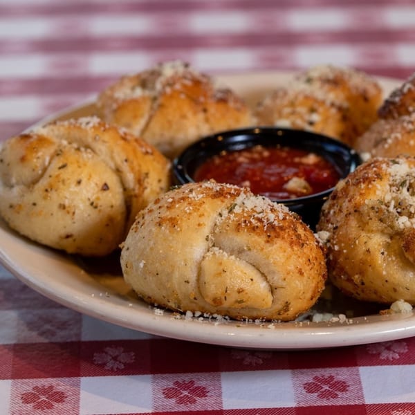 Garlic knots with parmesan and herbs, served with a side of marinara sauce on a red and white checkered tablecloth.