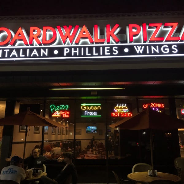 Exterior of Boardwalk Pizza at night, with neon signs and patrons dining inside and outside.