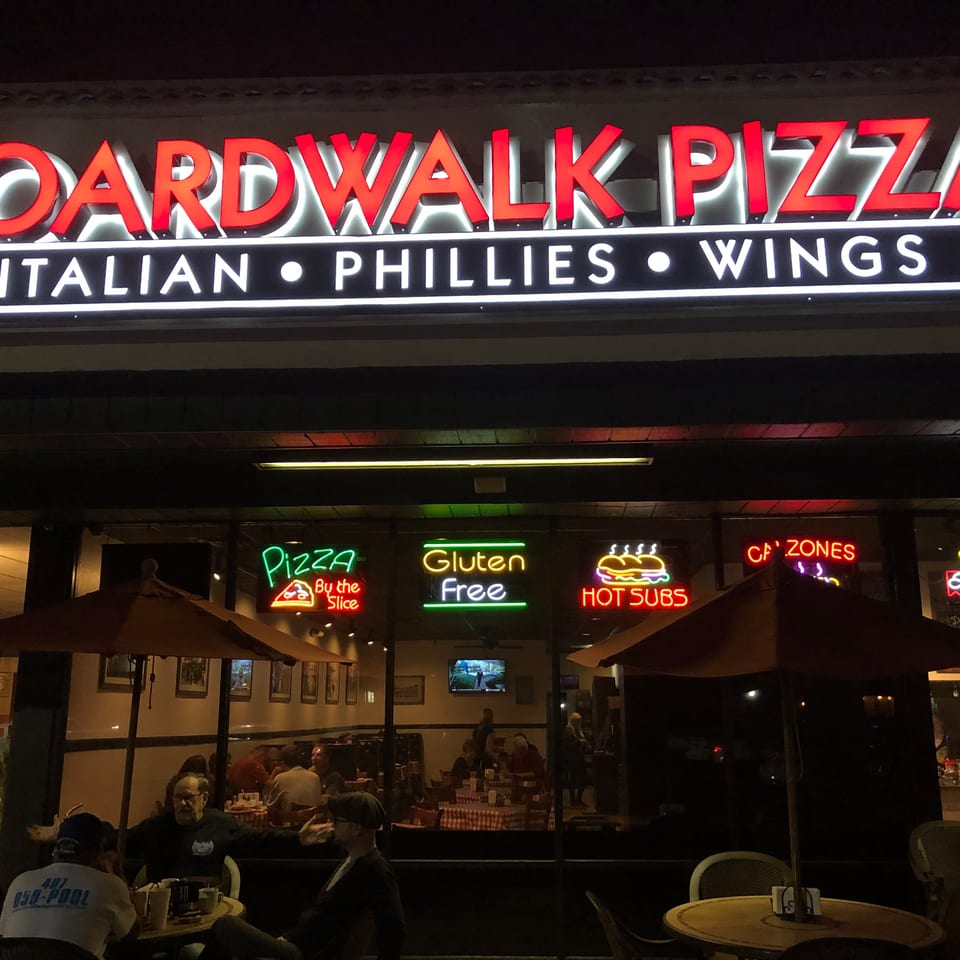 Exterior of Boardwalk Pizza at night, with neon signs and patrons dining inside and outside.