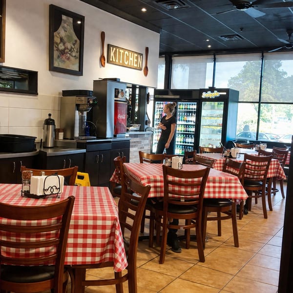 A restaurant dining room with red and white checkered tablecloths, a woman by drink coolers, and a "KITCHEN" sign.