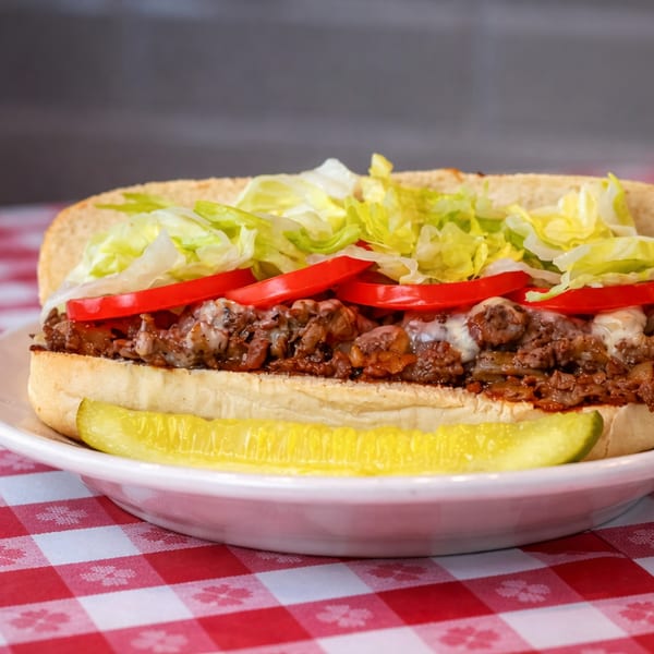 Cheesesteak sandwich with lettuce, tomatoes, and a pickle spear on a white plate with a red and white checkered tablecloth.
