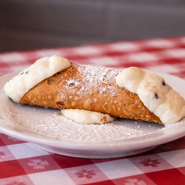 A cannoli with powdered sugar and cream filling, on a white plate over a red and white checkered tablecloth.