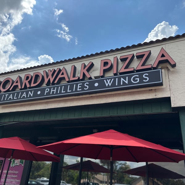 The Boardwalk Pizza sign with red umbrellas in front, under a blue sky with clouds.