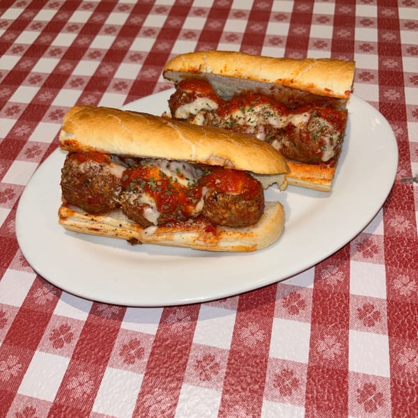 Two meatball subs with melted cheese and red sauce on a white plate, on a red and white checkered tablecloth.
