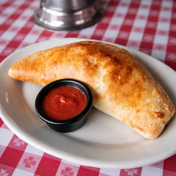 Golden-brown calzone with a side of marinara sauce on a white plate and red checkered tablecloth.