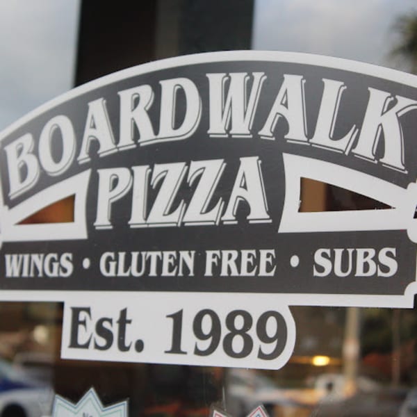 A close-up of the Boardwalk Pizza sign, featuring "Wings," "Gluten Free," "Subs," and "Est. 1989."